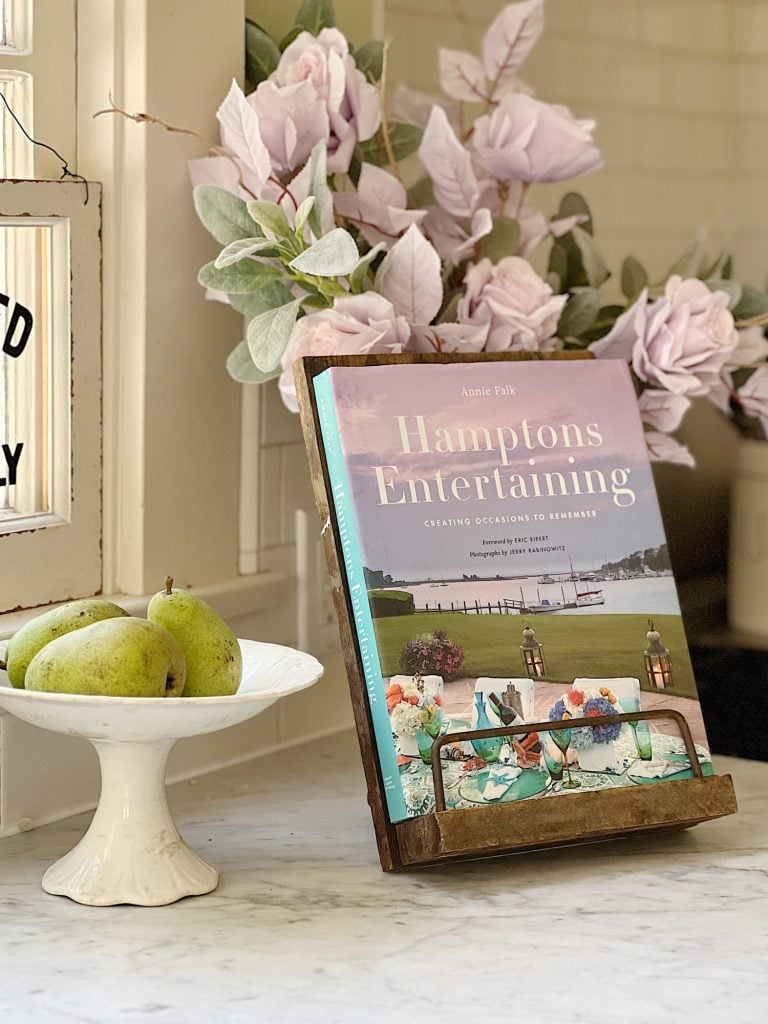 A book titled Hamptons Decorating, a white compote with green pears, a vase with faux lavender flowers, all sitting in a kitchen atop a white marble countertop.