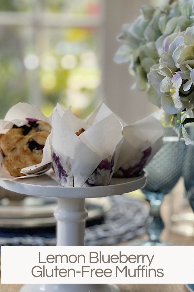 Lemon Blueberry Gluten-Free Muffins in white wrappers sitting on a cake stand next to a vase with light blue hydrangeas.
