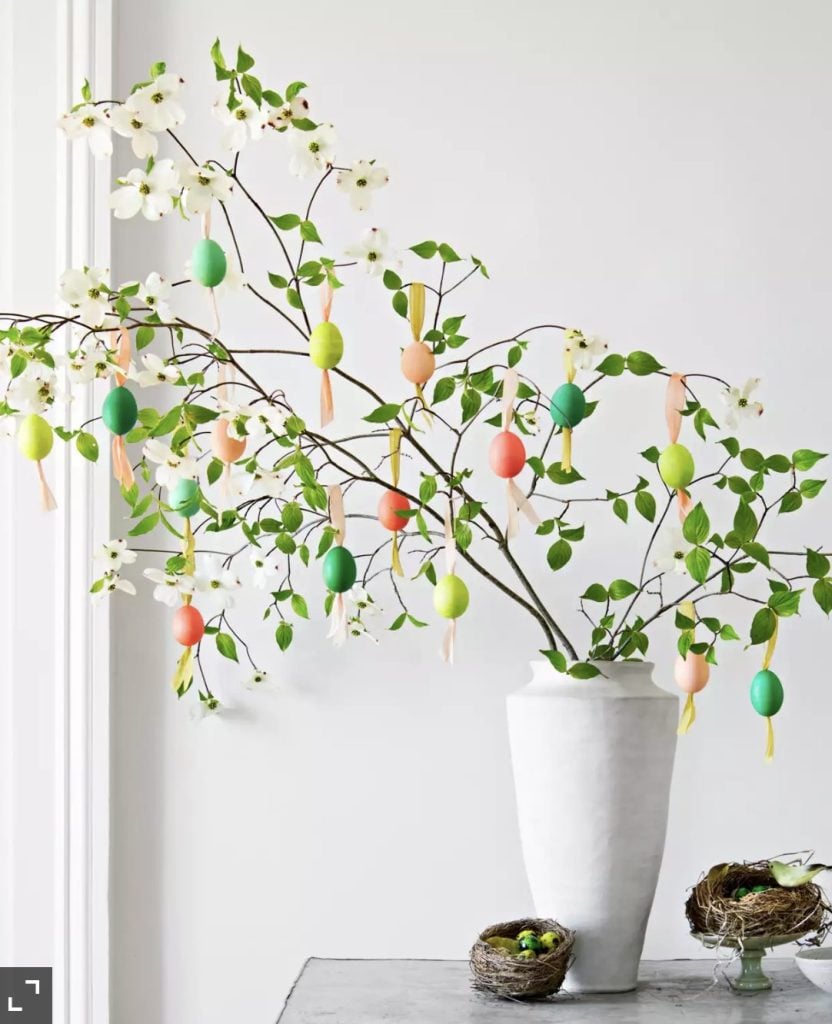 Tall what vase with branches. Hanging from the branches are colorlfully dyed easter eggs hanging from colorful ribbons