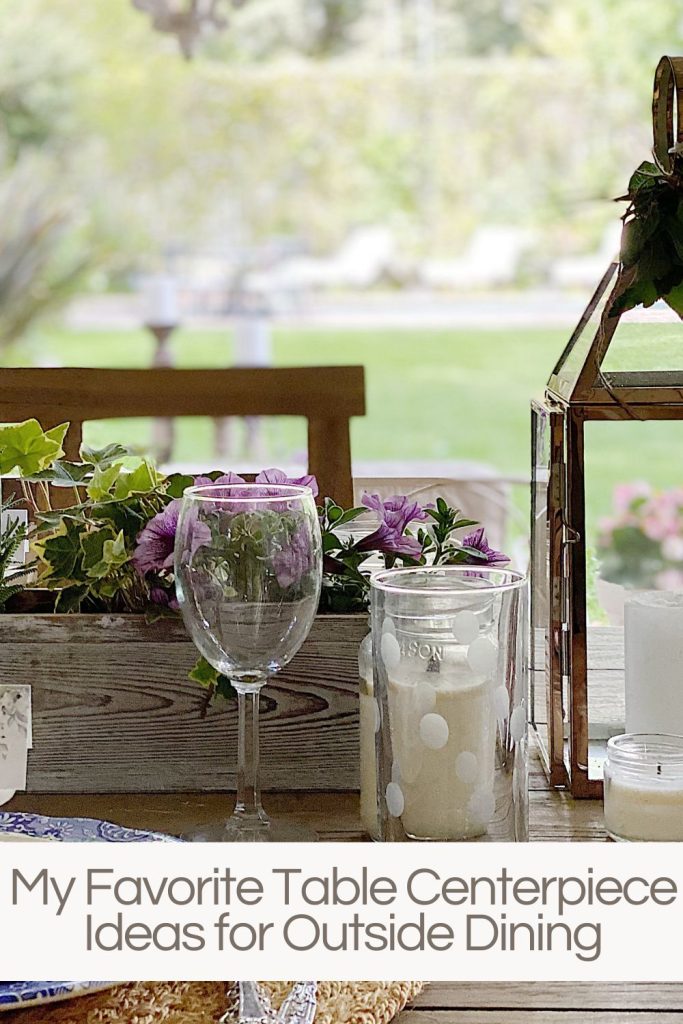 An outdoor table set for dinner with blue and white plates, polka dot glasses, copper glass candle lanterns, and centerpieces of white wood and fresh purple flowers.
