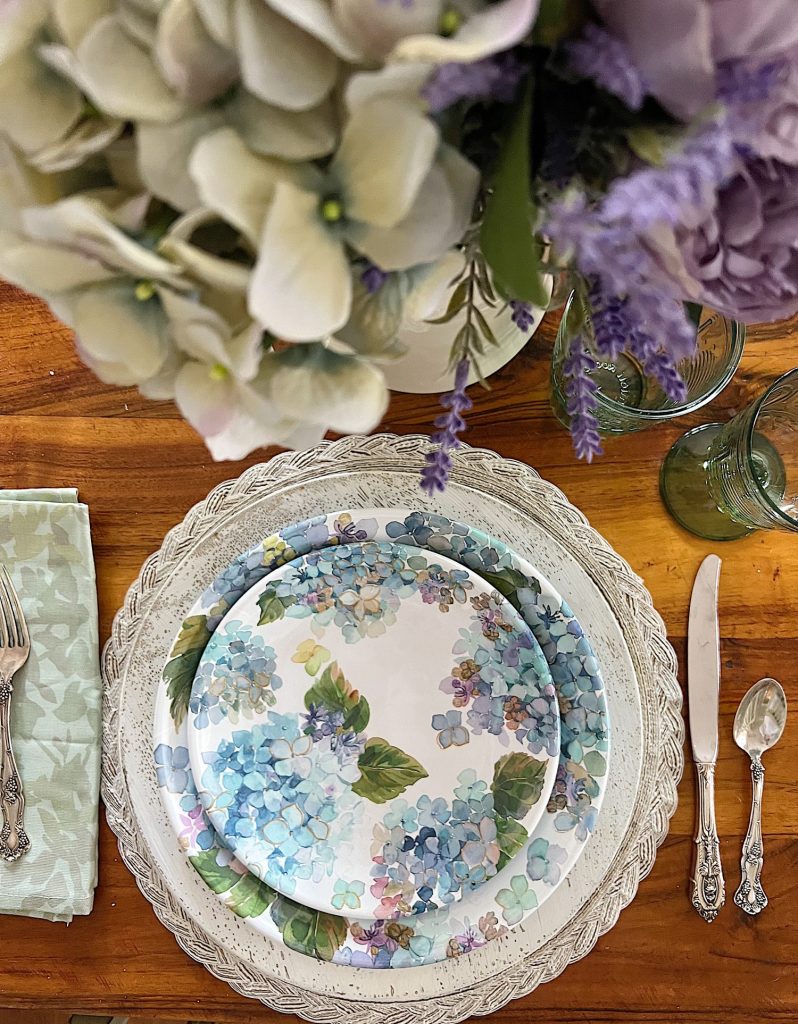 New spring home table in the family room with lavender and sage green flowers, hydrangea themed plates, and small decor items.