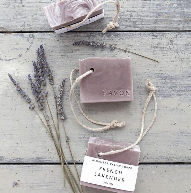 Three bars of handmade lavender scented soap, in french savon, each with a short rope. The soaps are on a white-washed table top with dried lavender scattered between the bars.