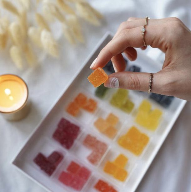 CBD gummies organized by color, 4 in each color group on a white tray. Candle and pampas grass in background. Hand with multiple gold rings holding an orange gummy at top of photo