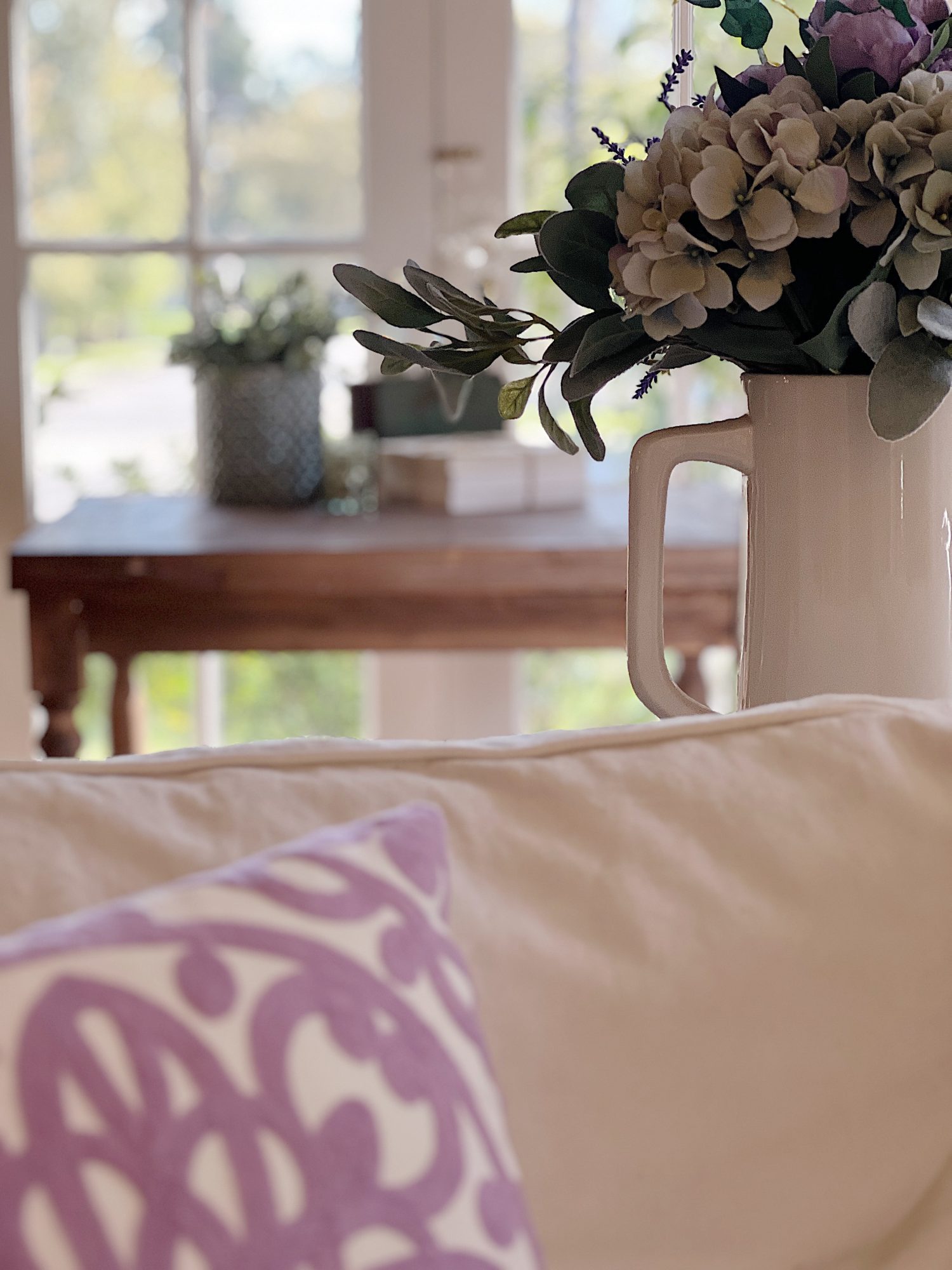A large living room with two white couches, a landscape painting above the mantle, sage green and lavender pillows, lavender throws, and spring decor.