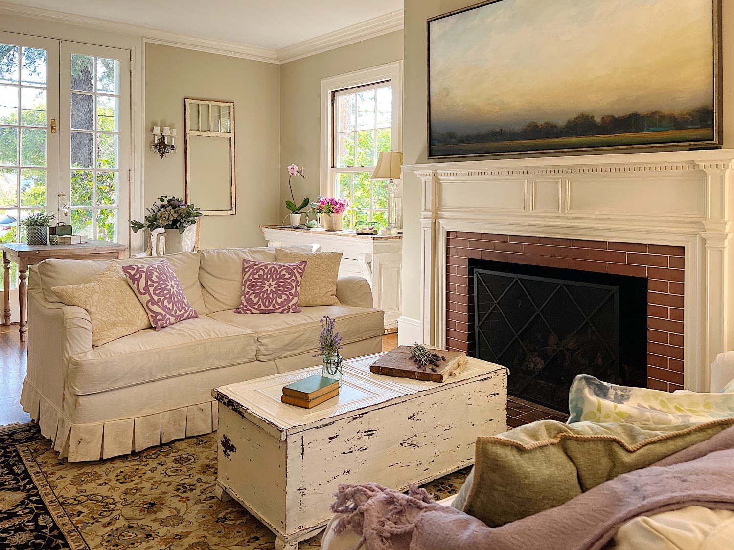 A large living room with two white couches, a landscape painting above the mantle, sage green and lavender pillows, lavender throws, and spring decor.