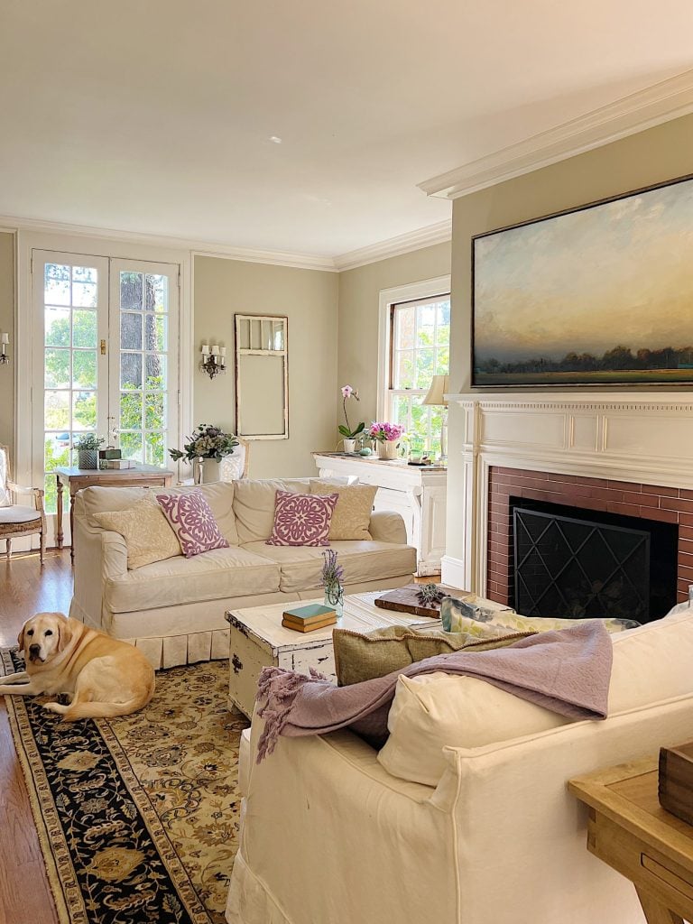 A large living room with two white couches, a landscape painting above the mantle, sage green and lavender pillows, lavender throws, and spring decor.