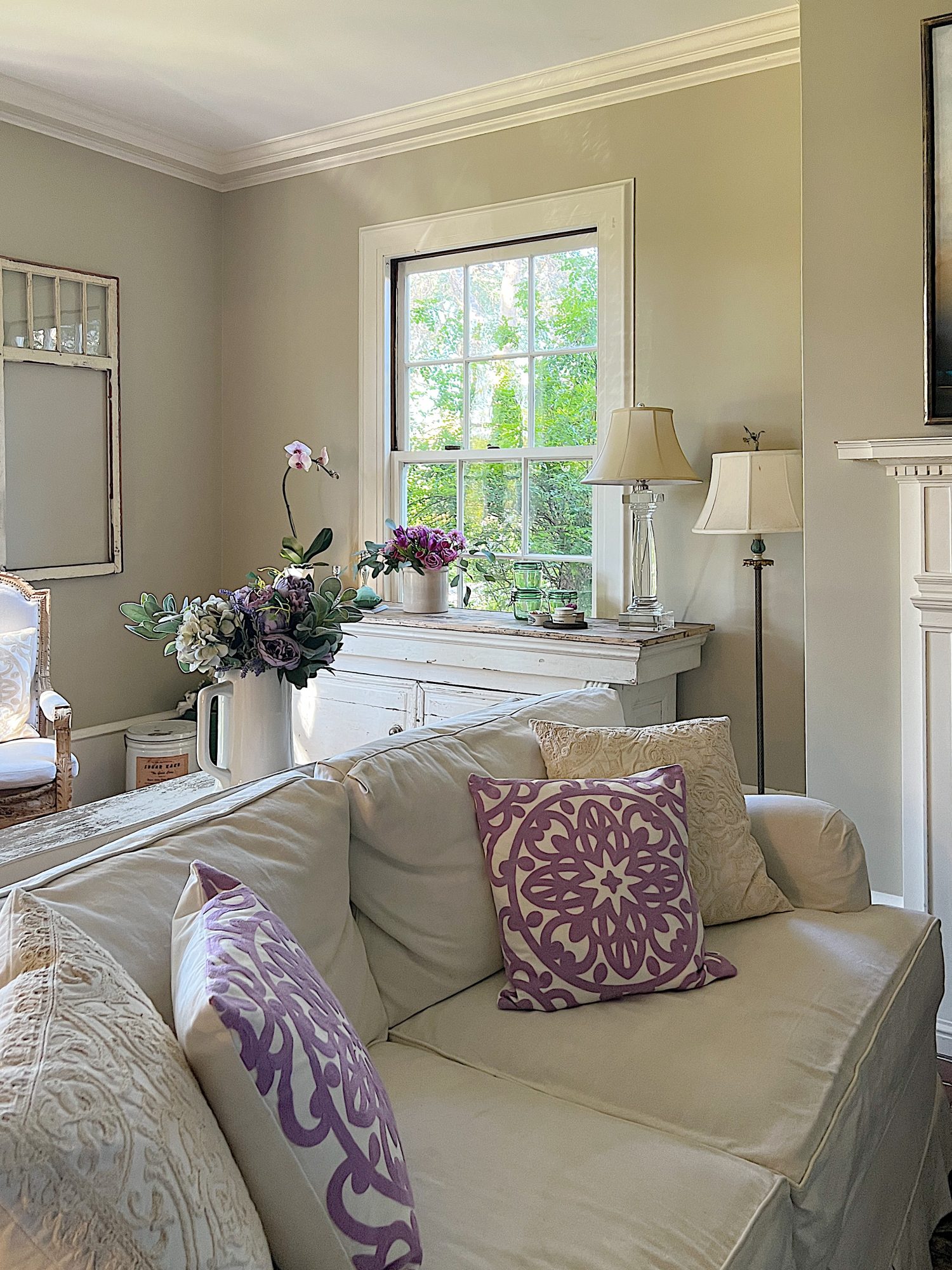 A large living room with two white couches, a landscape painting above the mantle, sage green and lavender pillows, lavender throws, and spring decor.
