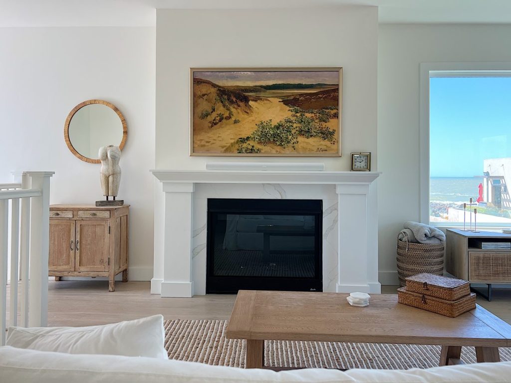 The living area at our beach house with a large fireplace, a frame tv, a window overlooking the ocean, and a couch, rug, coffee table and chair from Pottery Barn. The vintage cabinet, marble bust, and round rattan mirror are at the top of the staircase.
