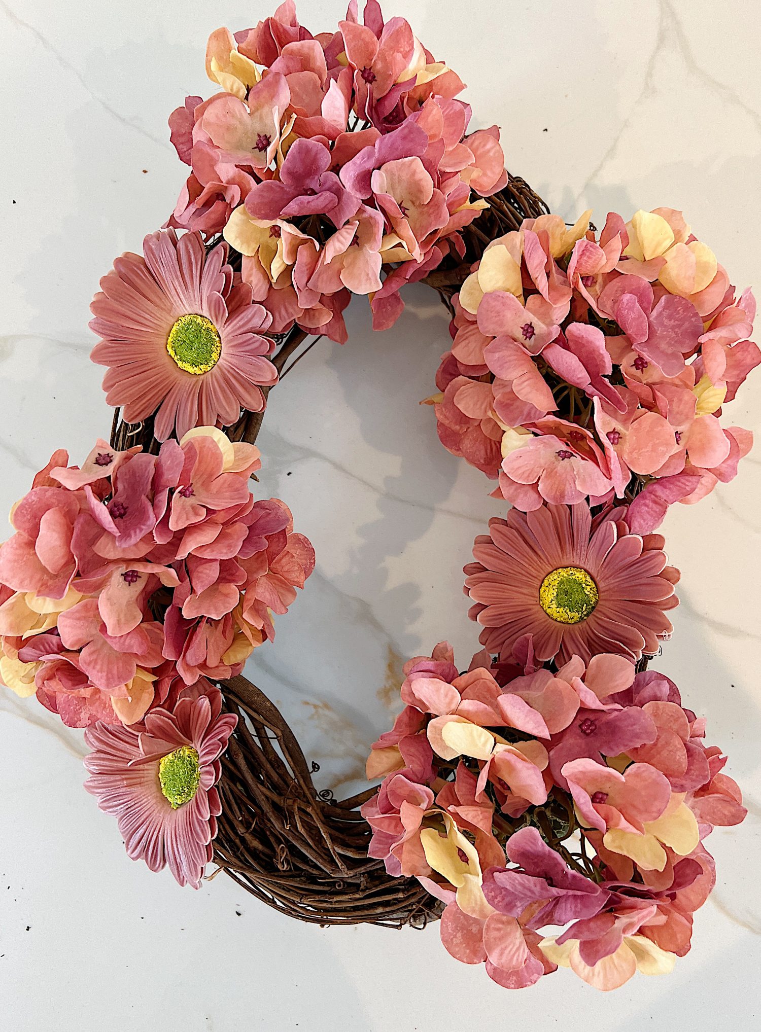 Handmade wreath consisting of faux hydrangeas, gerbera daisies, and roses attached to a grapevine wreath