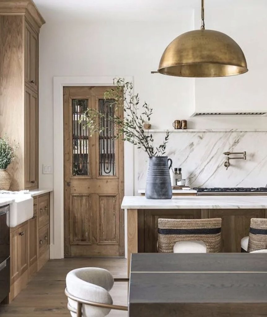 Kitchen with light walnut cabinets, an antique door with ironwork in the panels and a large brass dome light fixture which hangs over a kitchen osland. In the foreground is a dark wood table with cream upholstery and brass structure. The room is very calm and peaceful