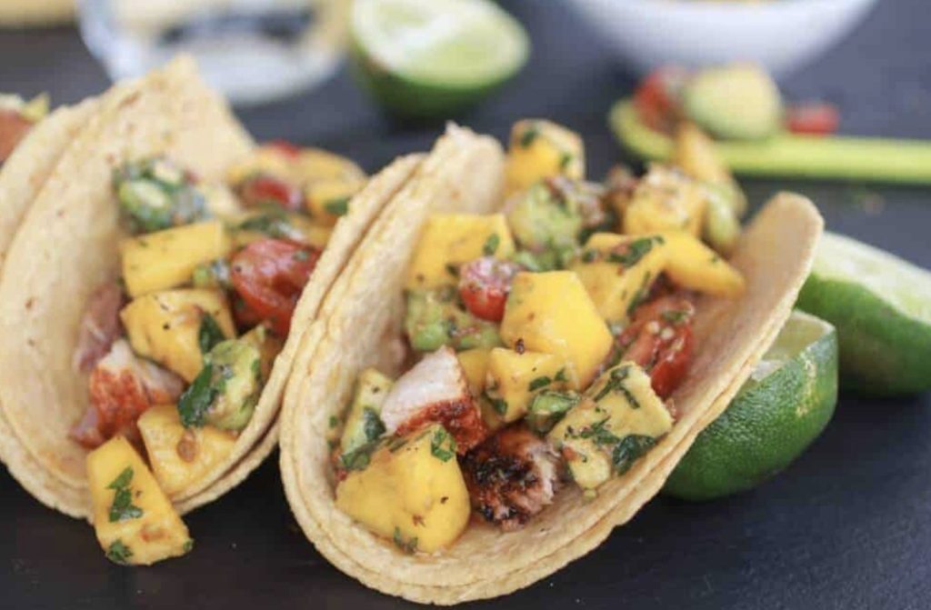 Two fish tacos packed with mango, fish, avocado, cilantro and tomatoes. Fresh squeezed limes are nearby