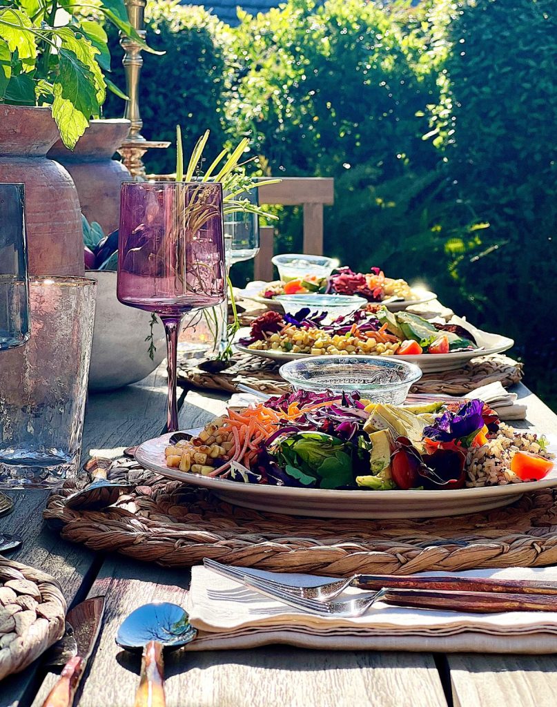 Earth Day dinner with Arhaus table, chairs, plates, colored glassware, salmon salad, copper silverware, and large stoneware vases with live tomato plants.