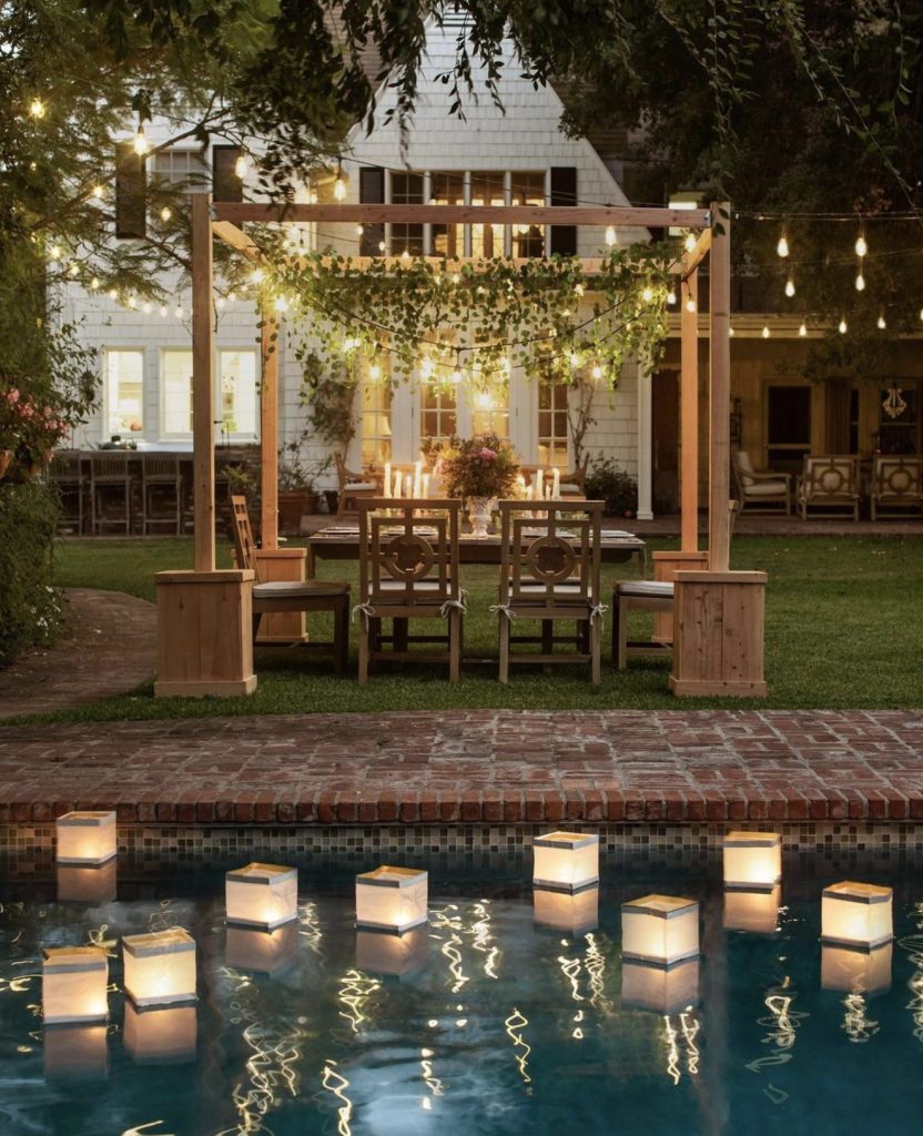 Yard with string lights strung through the trees and white house with lights on in background. In the foreground, floating lanterns reflect in the pool water. Inthe yard, a table is set amongst a wooden post structures. The table is set with festive flowers and many candles