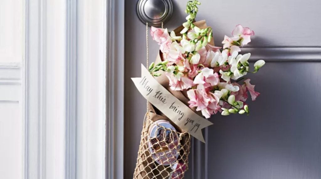 Fishermans net bag filled with sweets and goodies, with a bunch of pink snap drags tucked into the top hanging from a door knob on a gray front door. The net bag is embellished with a banner shaped piece of paper with black handwriting reading "May this bring you joy"