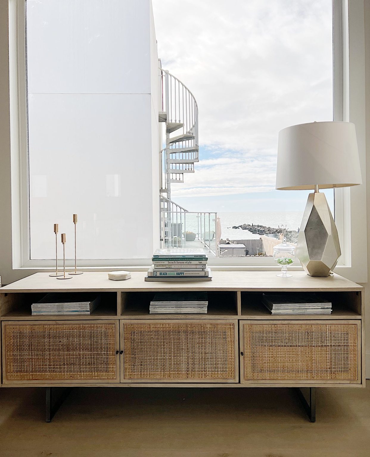 A cane media cabinet with decor and a modern gold lamp and a view of the ocean.