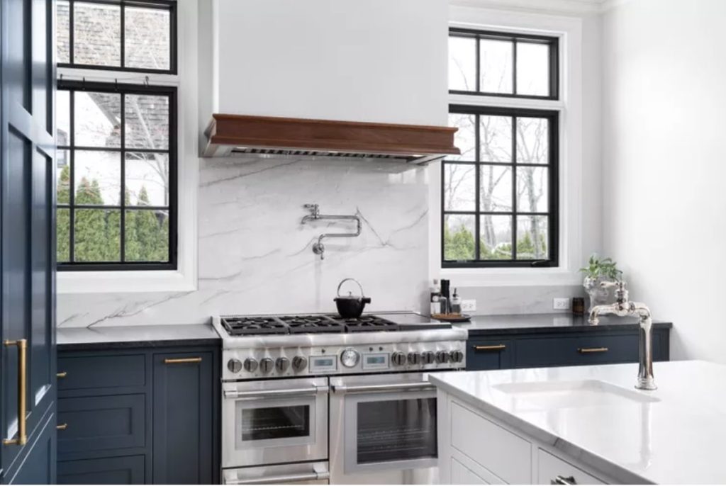 Beautiful kitchen with deep blue cabinetry, marble counters and stove backsplash and industrial windows. Stainless steel 8 burner stove with pot filler is at center of photo