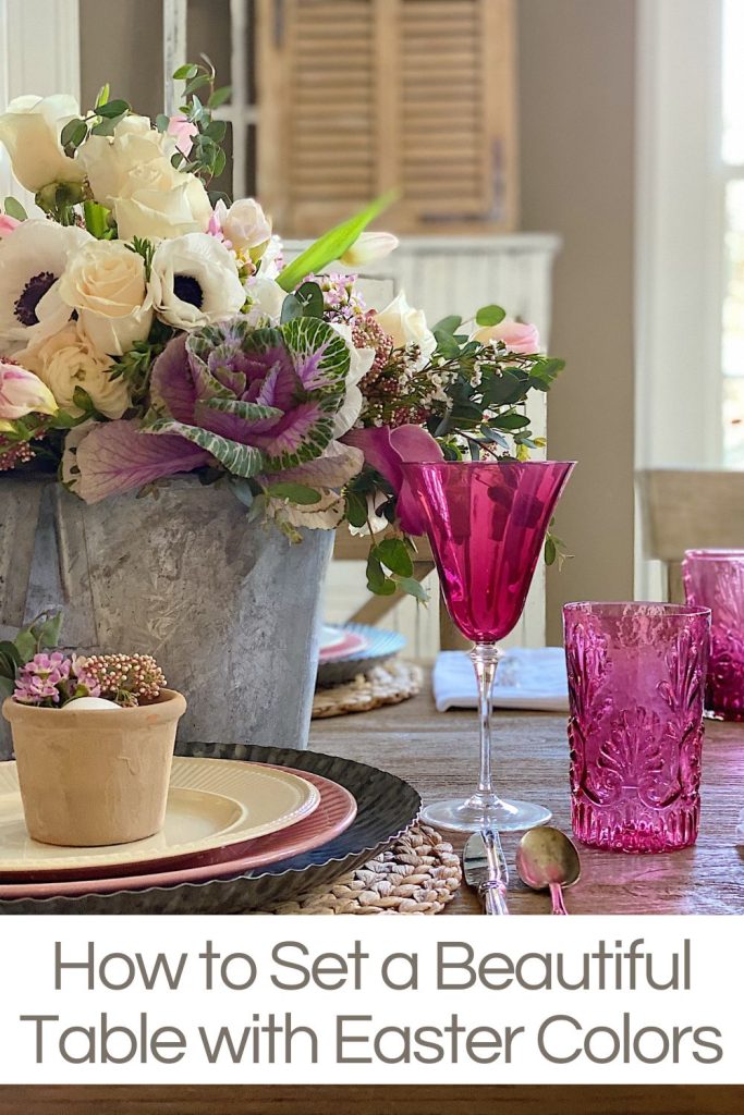 An Easter table with fresh flowers in a vintage calf bucket, terra cotta pots with flowers, and a wooden egg, china, and vintage cranberry glass.