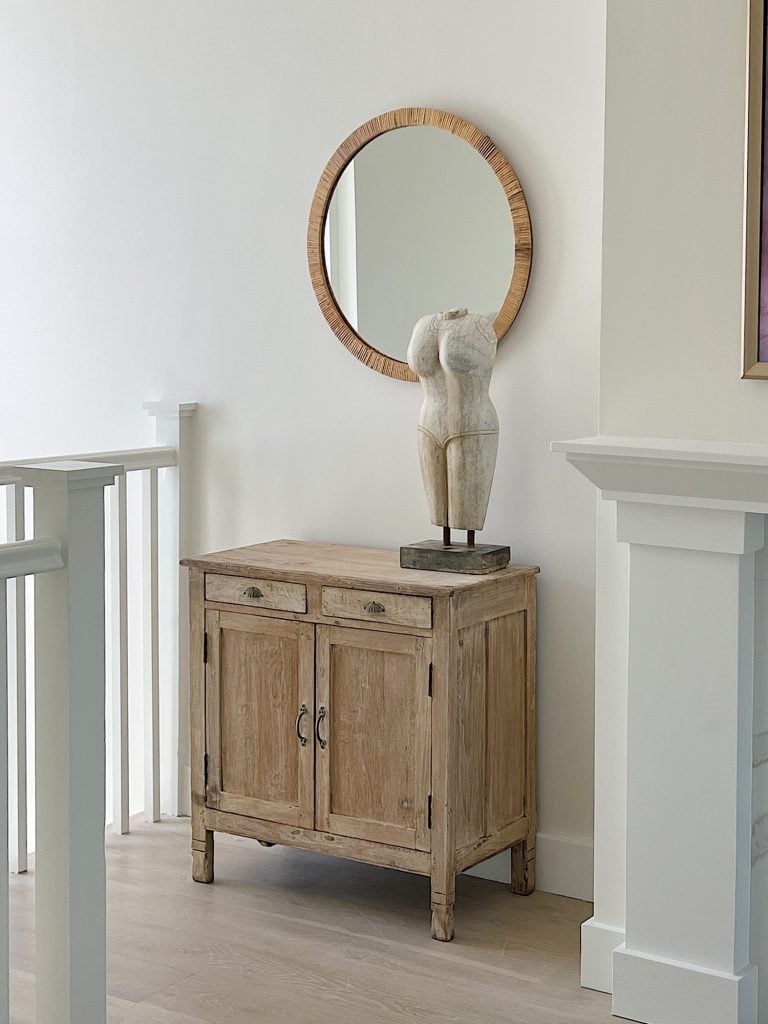 A white staircase with a vintage cabinet and a marble bust sitting on top and a round mirror on the wall.