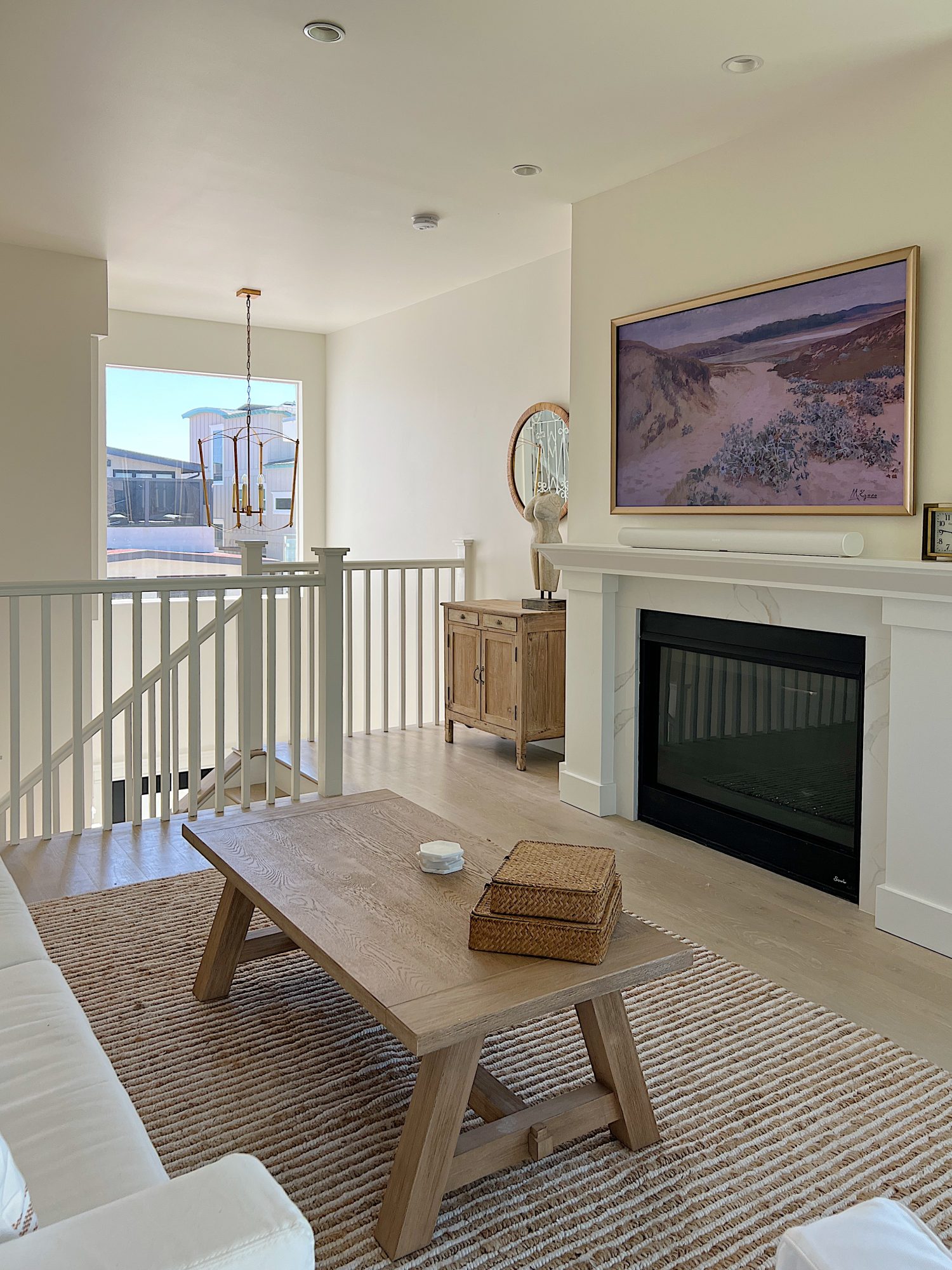 The living area at our beach house with a large fireplace, a frame tv, a window overlooking the ocean, and a couch, rug, coffee table and chair from Pottery Barn. The vintage cabinet, marble bust, and round rattan mirror are at the top of the staircase.