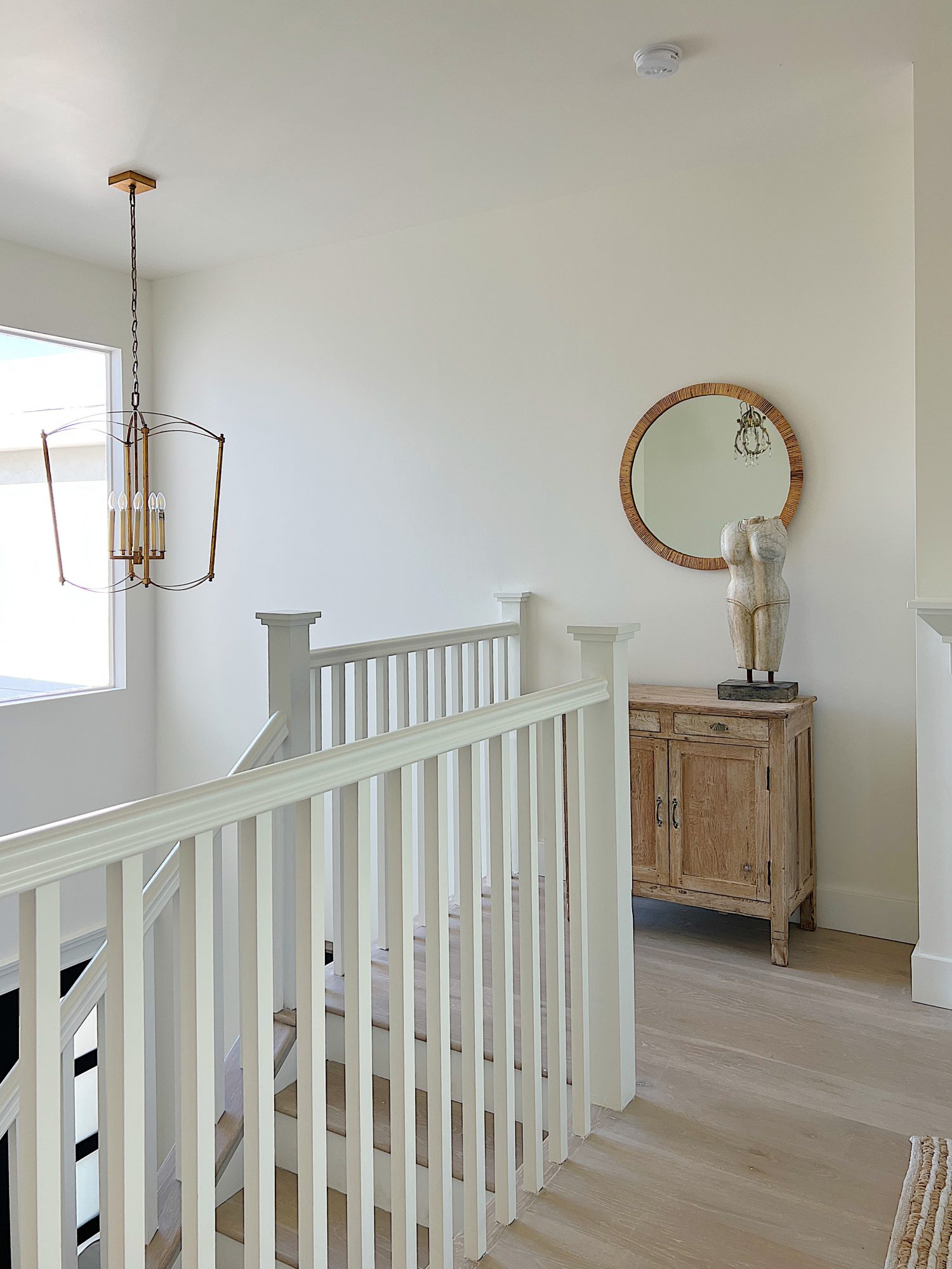 A white staircase with a vintage cabinet and a marble bust sitting on top and a round mirror on the wall.