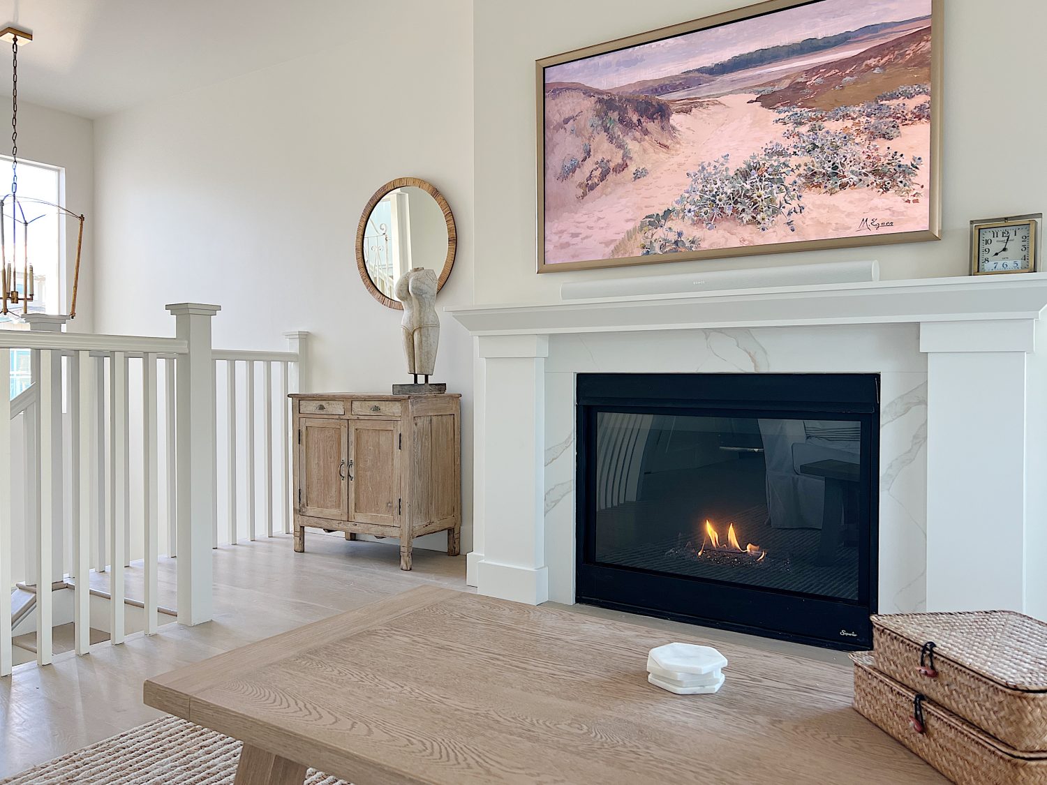 The living area at our beach house with a large fireplace, a frame tv, a window overlooking the ocean, and a couch, rug, coffee table and chair from Pottery Barn. The vintage cabinet, marble bust, and round rattan mirror are at the top of the staircase.