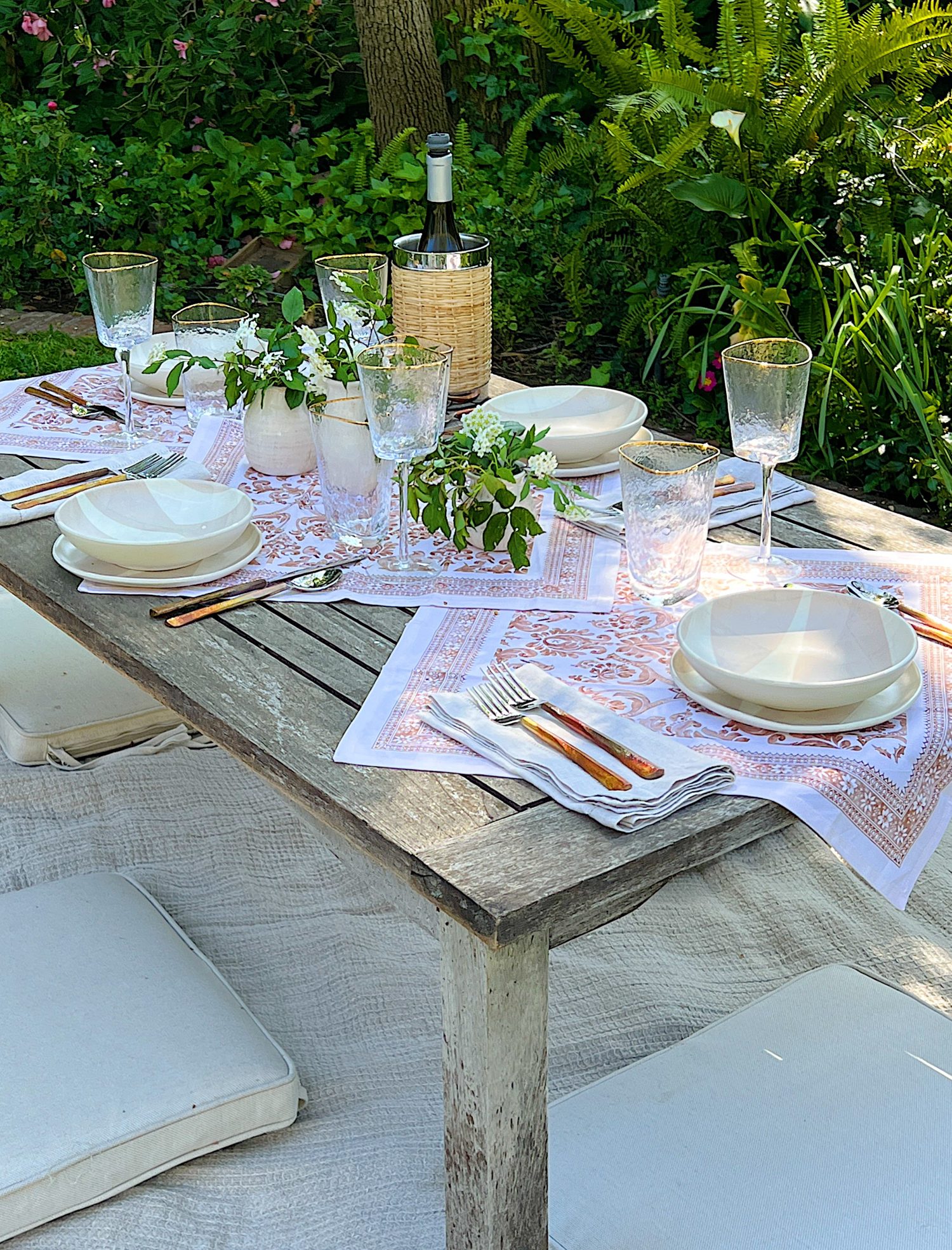 A picnic set up on a blanket that includes plastic dishes, glassware, copper forks, vases and fresh flowers, and rattan barware.