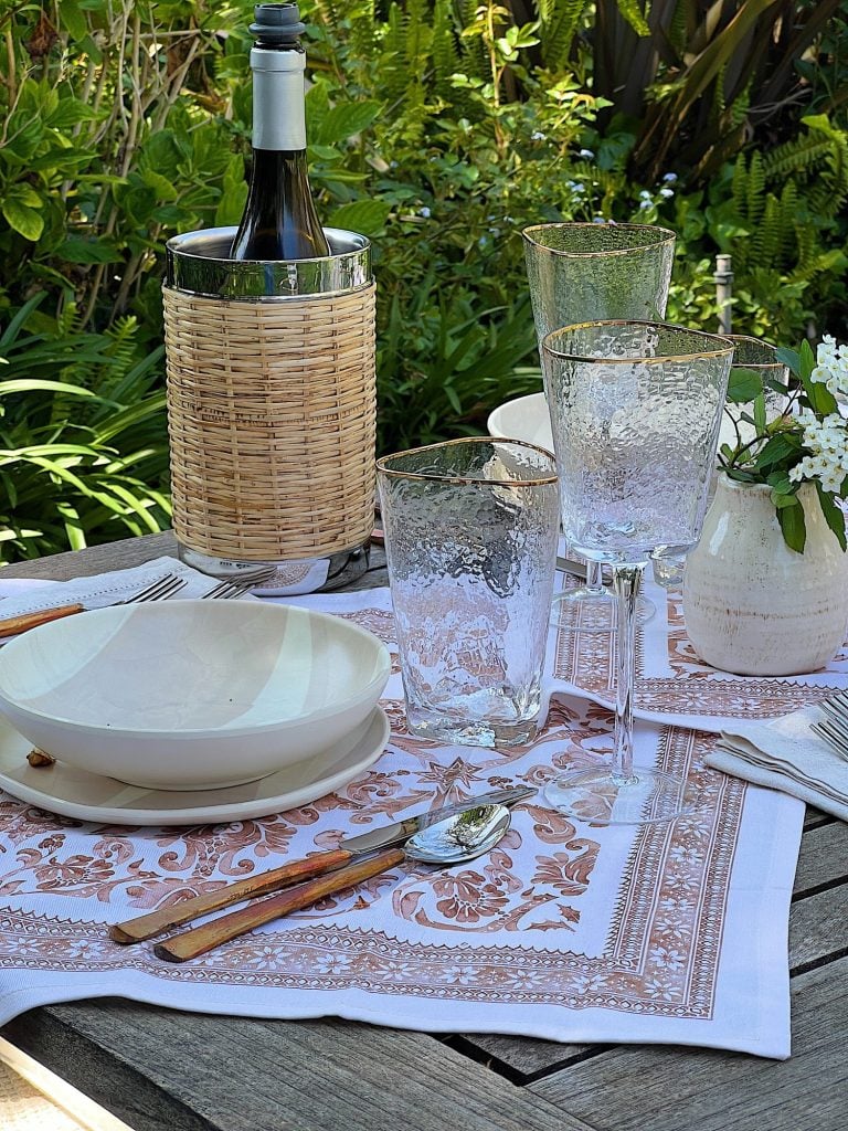 A picnic set up on a blanket that includes plastic dishes, glassware, copper forks, vases and fresh flowers, and rattan barware.