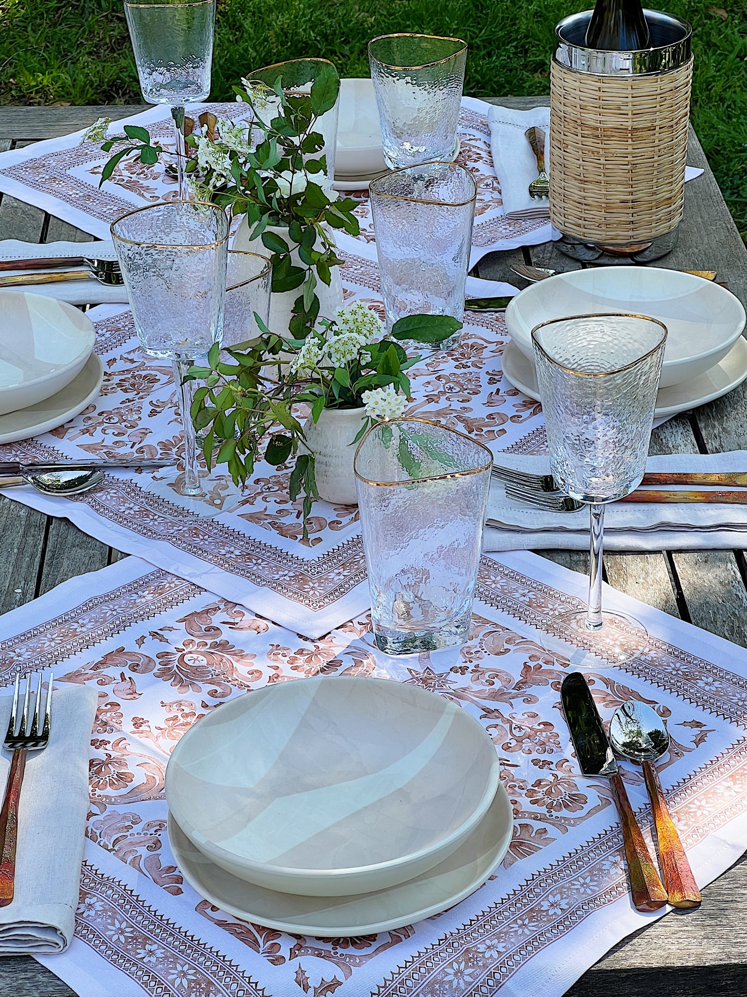 A picnic set up on a blanket that includes plastic dishes, glassware, copper forks, vases and fresh flowers, and rattan barware.