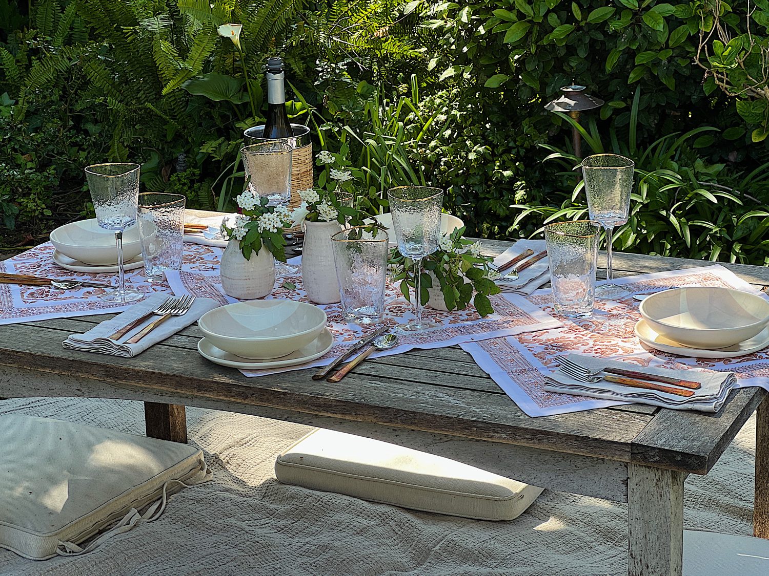 A picnic set up on a blanket that includes plastic dishes, glassware, copper forks, vases and fresh flowers, and rattan barware.