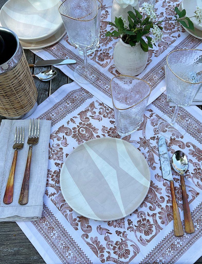 A picnic set up on a blanket that includes plastic dishes, glassware, copper forks, vases and fresh flowers, and rattan barware.