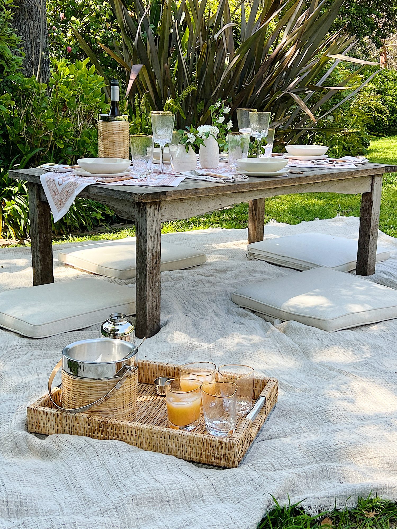 A picnic set up on a blanket that includes plastic dishes, glassware, copper forks, vases and fresh flowers, and rattan barware.