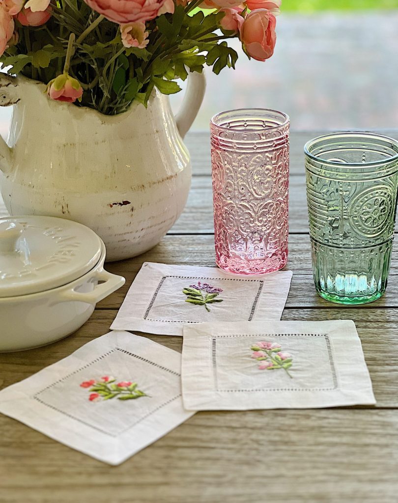Cotton/linen napkins embroidered with flowers and colored glassware, a brie maker, and a white vase with faux flowers.