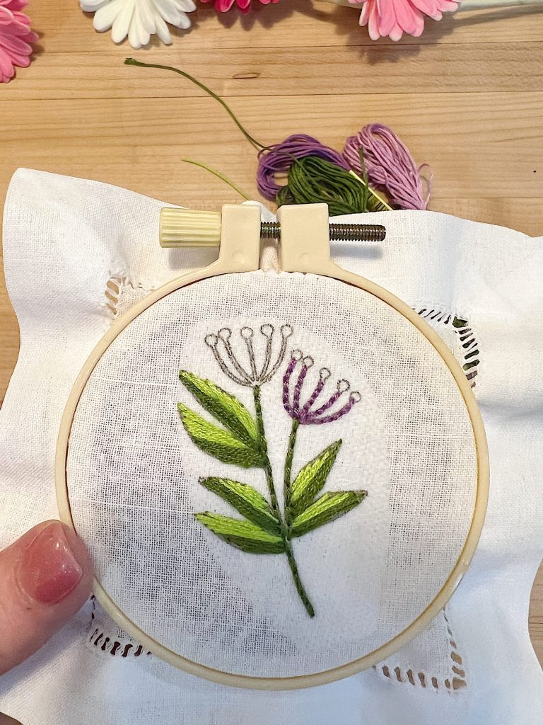 A cotton and linen hemstitched cocktail napkin, a stick-and-stitch floral embroidery pattern, an embroidery hoop, and embroidery thread.