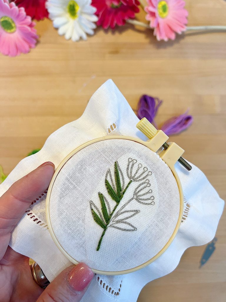 A cotton and linen hemstitched cocktail napkin, a stick-and-stitch floral embroidery pattern, an embroidery hoop, and embroidery thread.