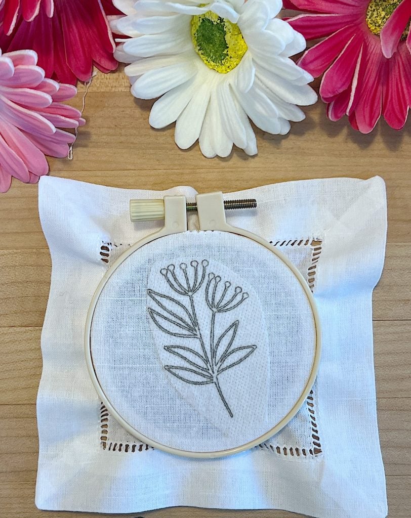A cotton and linen hemstitched cocktail napkin, a stick-and-stitch floral embroidery pattern, an embroidery hoop and faux flowers.