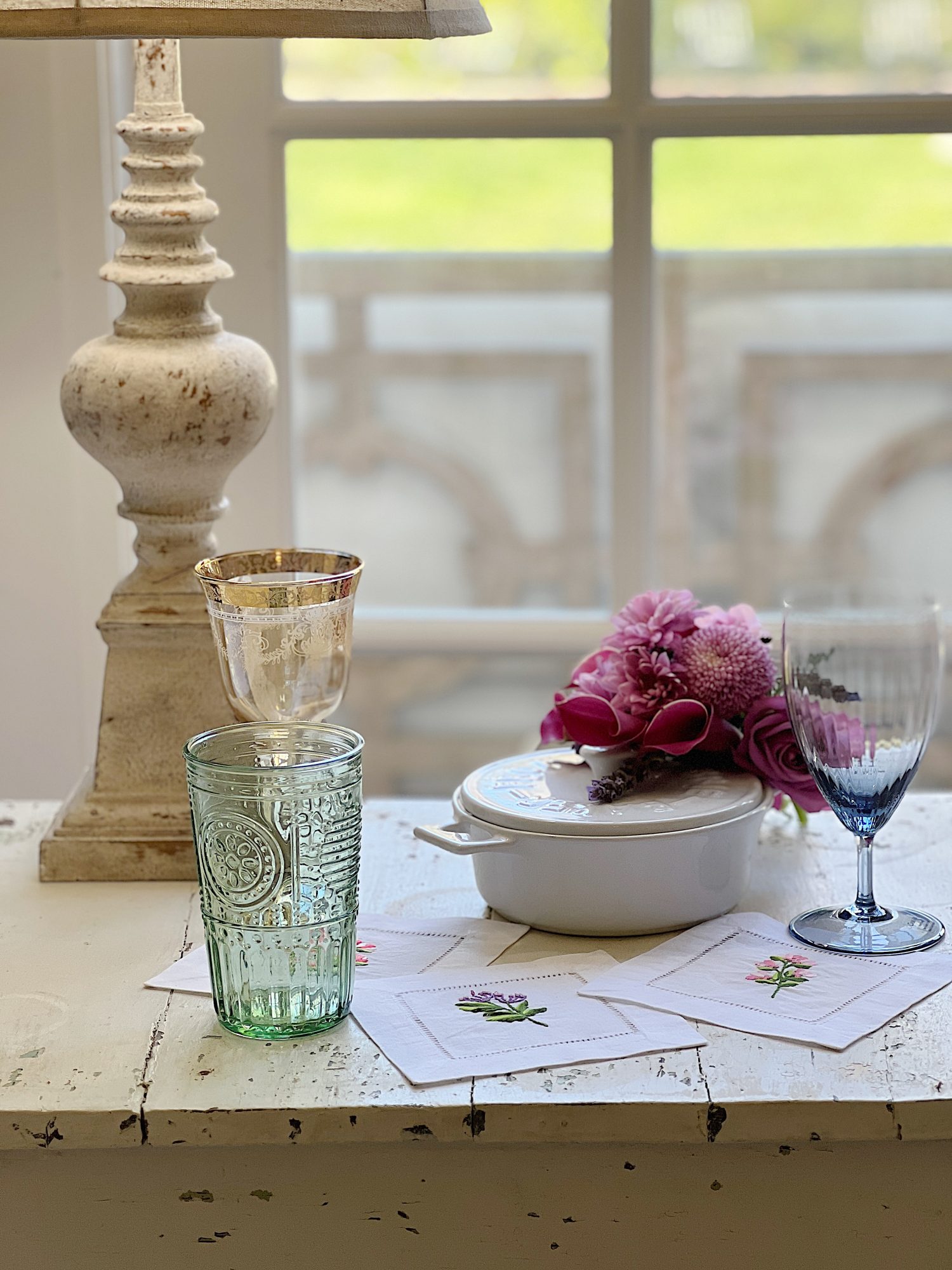 Cotton/linen napkins embroidered with flowers and colored glassware, a brie maker, and a white vase with faux flowers.