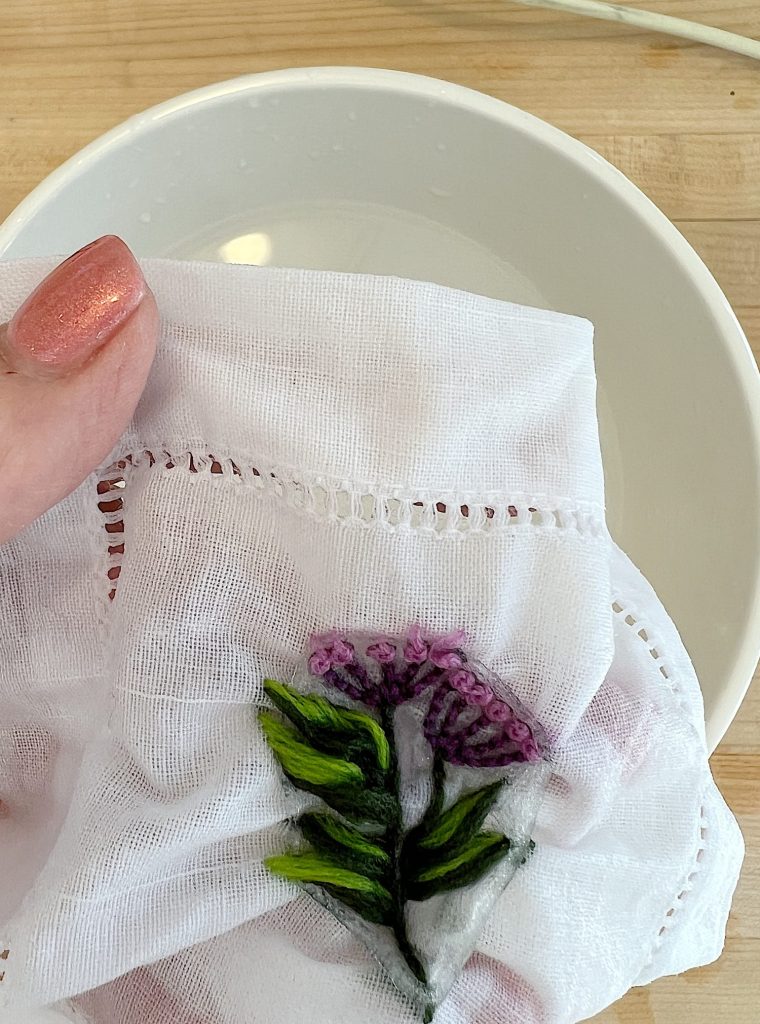 A cotton and linen hemstitched cocktail napkin embroidered using a stick-and-stitch floral embroidery pattern, and purple and green embroidery thread. It is in water to remove the pattern.