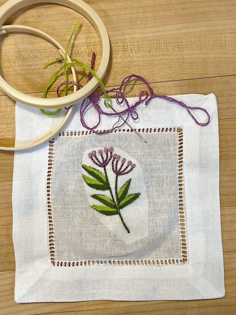 A cotton and linen hemstitched cocktail napkin embroidered using a stick-and-stitch floral embroidery pattern, and purple and green embroidery thread.