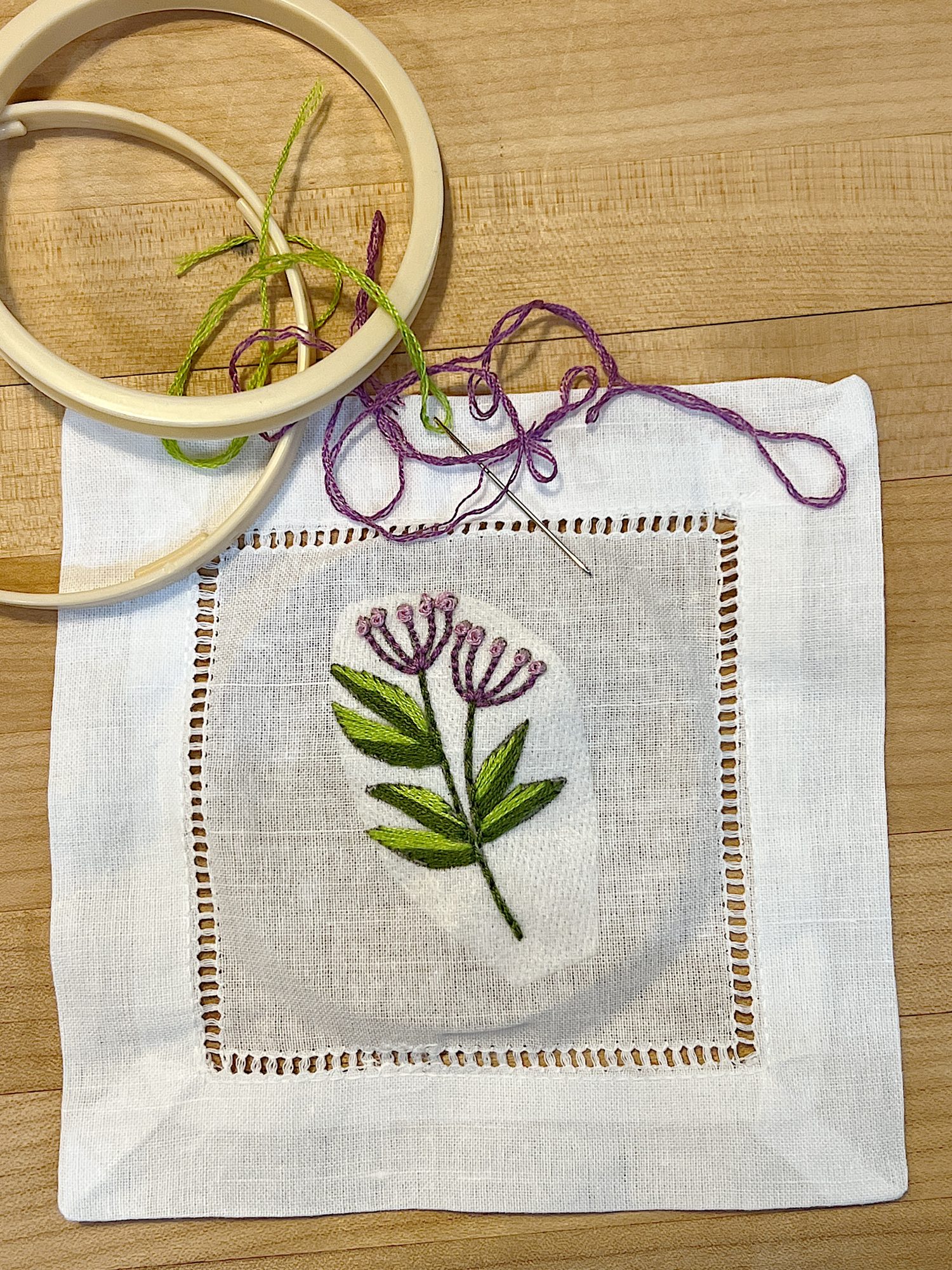 A cotton and linen hemstitched cocktail napkin embroidered using a stick-and-stitch floral embroidery pattern, and purple and green embroidery thread.