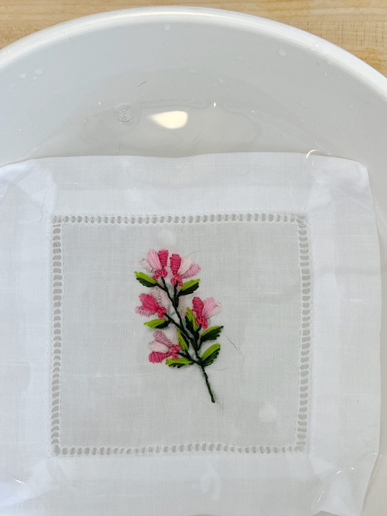 A cotton and linen hemstitched cocktail napkin embroidered using a stick-and-stitch floral embroidery pattern, and pink and green embroidery thread.
