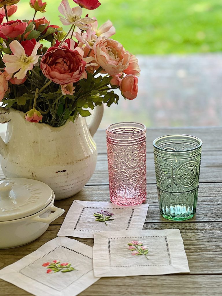 Cotton/linen napkins embroidered with flowers and colored glassware, a brie maker, and a white vase with faux flowers.