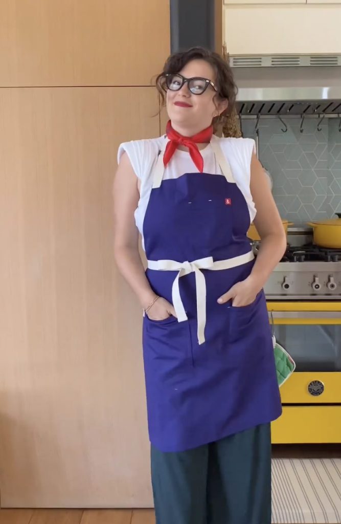 Beautiful woman in white t-shirt, a red kerchief tied around her neck wearing a blue Hedley & Bennett apron. A yellow range with yellow pots on top is in the background