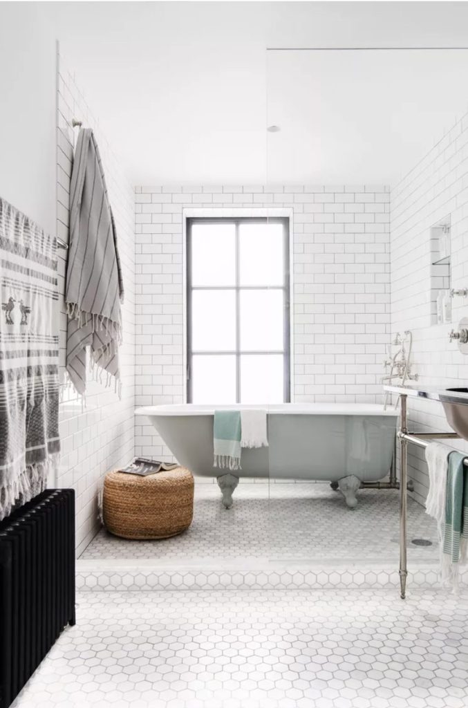 White and gray bathroom with subway tiles on wall and hexagon tiles on floor. Gray tub with towels draped over the edge near an industrial window, a rattan poof with books on top is next to the tub. Foreground is a peek at the sink on the right and a black radiator on the left. A gray and white striped towel hangs from a hook on the wall.