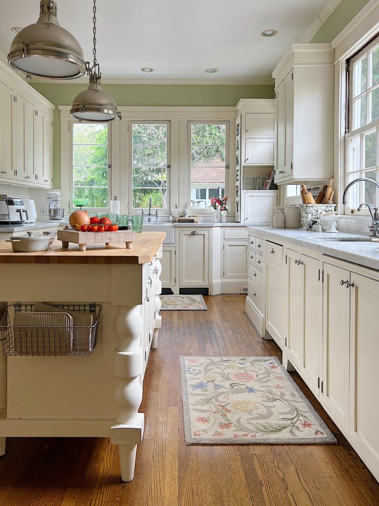 A white farmhouse kitchen with white shaker cabinets. honed marble countertops, an island with a butcher block top, and lots of farmhouse kitchen decor items.
