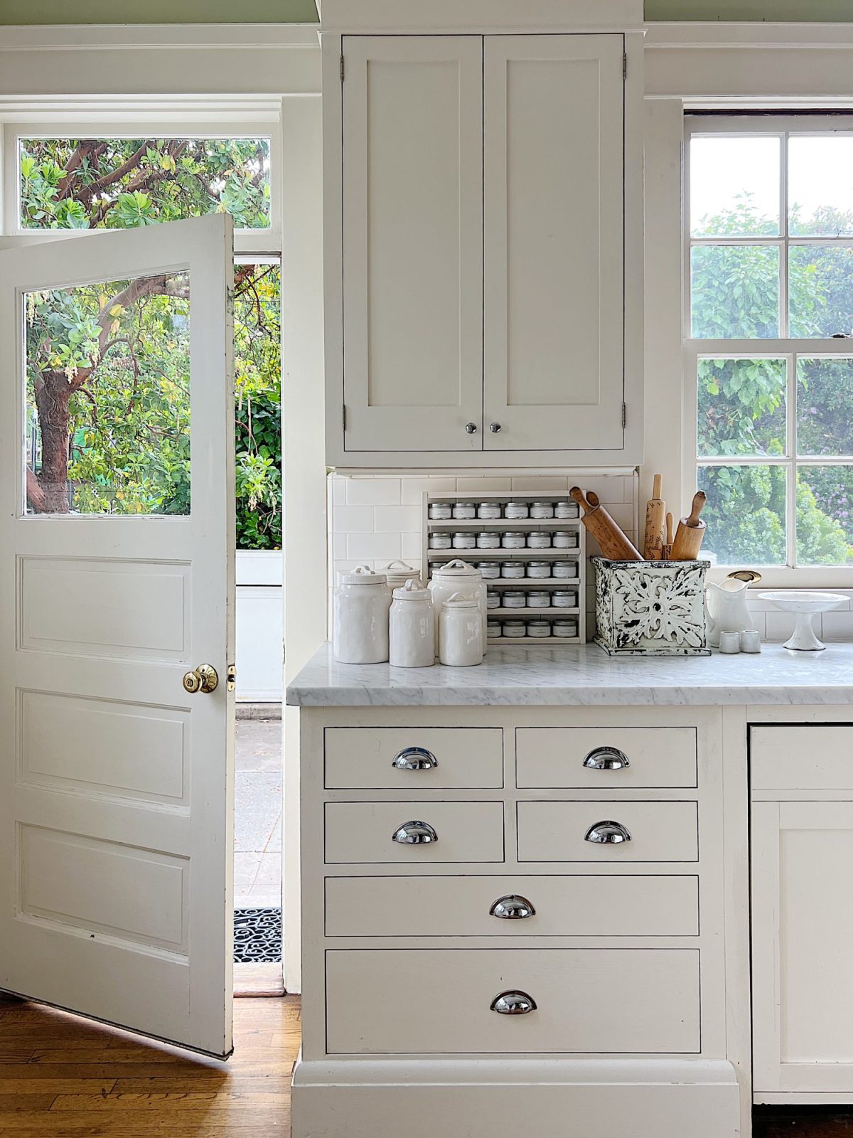A white farmhouse kitchen with white shaker cabinets. honed marble countertops, an island with a butcher block top, and lots of farmhouse kitchen decor items.