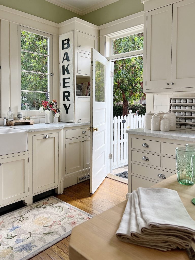 A white farmhouse kitchen with white shaker cabinets. honed marble countertops, an island with a butcher block top, and lots of farmhouse kitchen decor items.