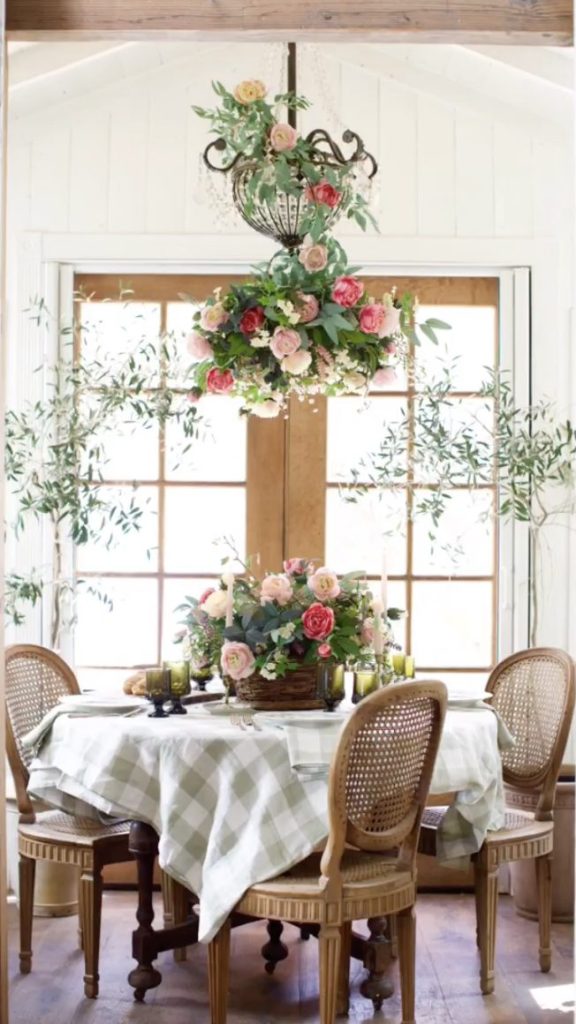 A French country table with sage and white buffalo plaid table linen is set in front of a French door. There is a large floral arrangement with large roses in various shades of pink, peach, and cream. Another flower arrangement hangs from the chandelier echoing the arrangement on the table.