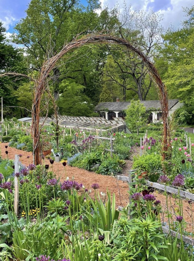 A beautiful potager garden with a home in the background. The flower garden is framed with an arch made of twigs while the vegetable garden is in the background.