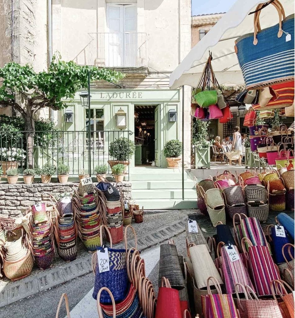 A street in Provence, France with a storefront painted in mint green, people are sitting at a table at an outdoor cafe in the background and colorful french market bags are lined beneath the steps of the shop