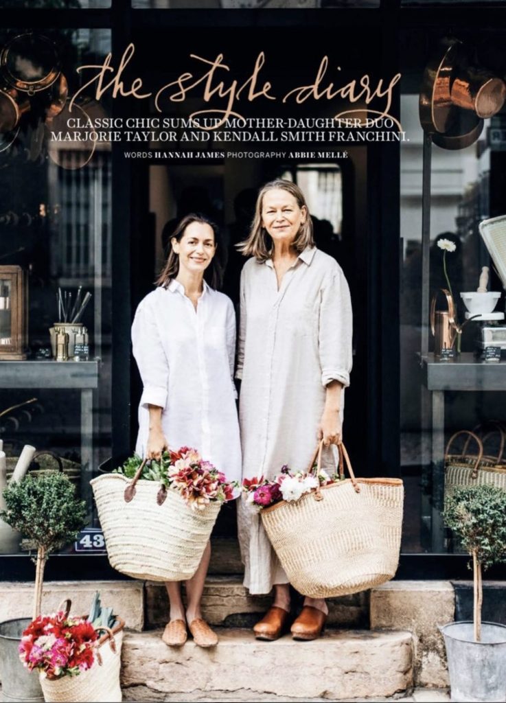 Two beautiful women standing on the limestone steps at the doorway of a storefront in France. they each hold a french market filled with flowers. They wear french linen and clogs.